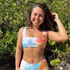 Woman wearing a sporty, colorful tie-dye swimsuit in swim shorts standing in a natural setting with greenery.