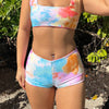 Woman wearing a sporty, colorful tie-dye swimsuit in swim shorts standing in a natural setting with greenery.