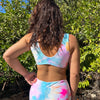 Woman wearing a sporty, colorful tie-dye swimsuit in swim shorts standing in a natural setting with greenery.
